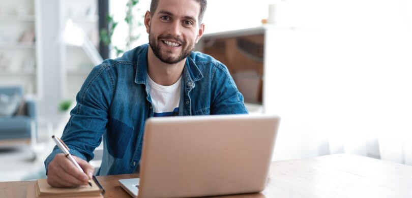 A man sits at a laptop computer in a modern, brightly lit office setting.